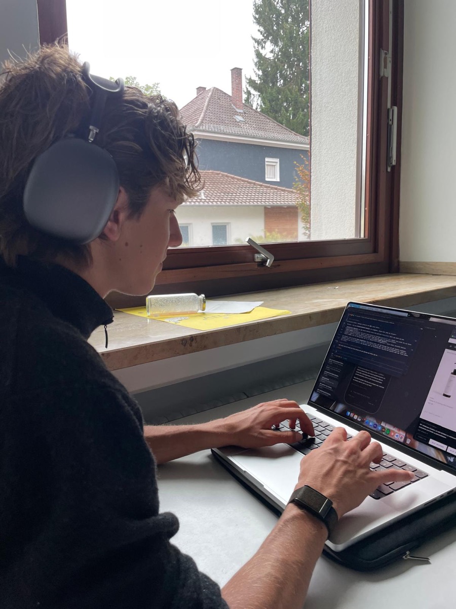 Elias Conrad im Profil bei der Arbeit am MacBook am Fenster, mit Kopfh&ouml;rern